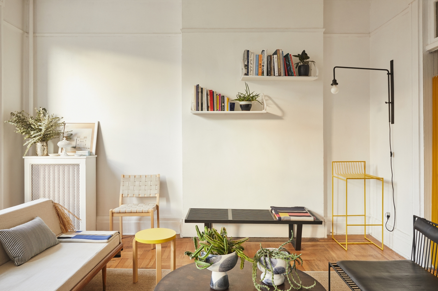 A pair of Alvar Aalto 112 Shelves is mounted above a vintage black George Nelson Platform Bench (a blanket of Valentin’s own design is folded on top of the bench). To the right of the bench is a yellow Bogus Studio Arnold Bar Chair and a wall-mounted swing arm lamp from Rejuvenation (no longer available) mounted above; Valentin removed the shade so the lamp would more closely resemble the Jean Prouvé Potence Lamp.