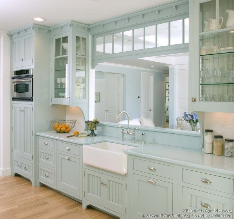A pale blue Victorian kitchen with matching blue countertops, a white apron sink, glass cabinets, and a large pass through window
